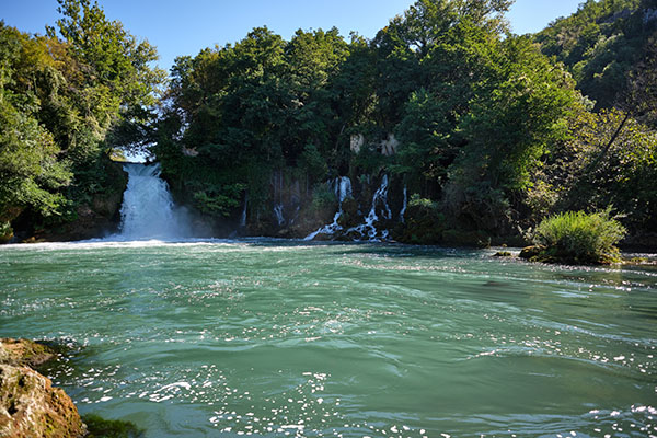Blick von unten auf einen breiteren und mehrere kleine Wasserfälle entlang von einem hufeisenförmigen stark bewachsenen Felsen; die Wasserfälle sind im Schatten, der daraus entstandene breite Fluss ist in der Sonne und wunderschön türkisfarben