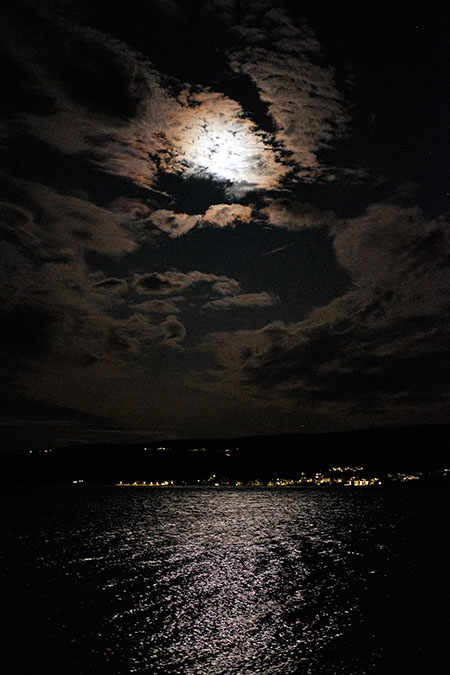 Mond hinter den Wolken, ein paar Lichter am gegenüberliegenden Meeresufer, das Mondlicht spiegelt sich im Meer