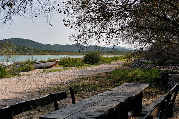 Vordergrund: Holzbänke und Holztisch, daneben ein Baum, dahinter ein Schotterweg und Wiese, weiter hinten ein See mit Schilf und Büschen am Ufer aus dem kleine Boote herausragen, hinter dem See grüne Hügeln